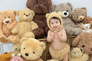 たくさんのクマの縫いぐるみの中、赤ちゃんがクマの着ぐるみを着ている